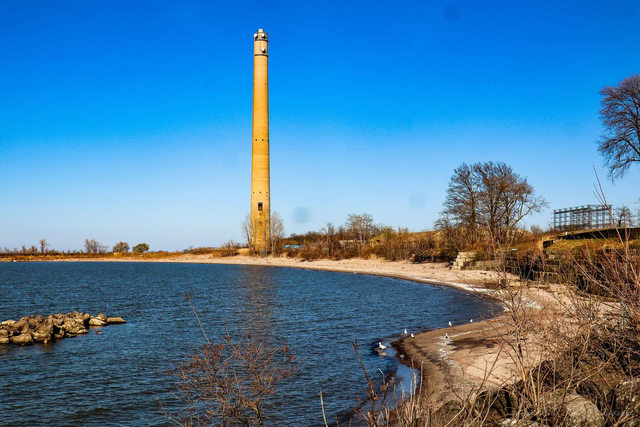 A tall disused smokestack above a curving sandy beach and blue water.