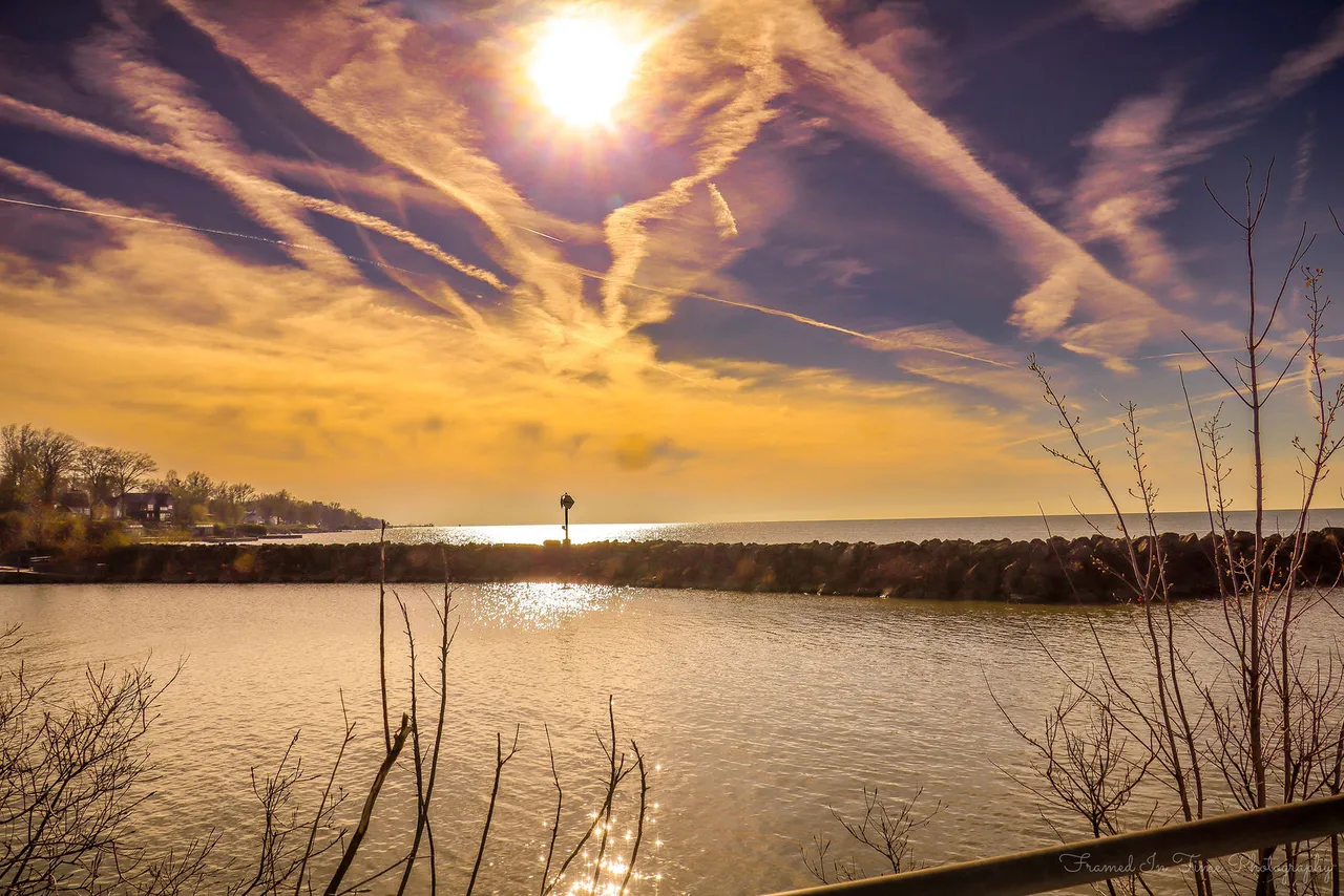 Golden sunlight over a rock breakwater under streaked clouds, bare branches in the foreground.