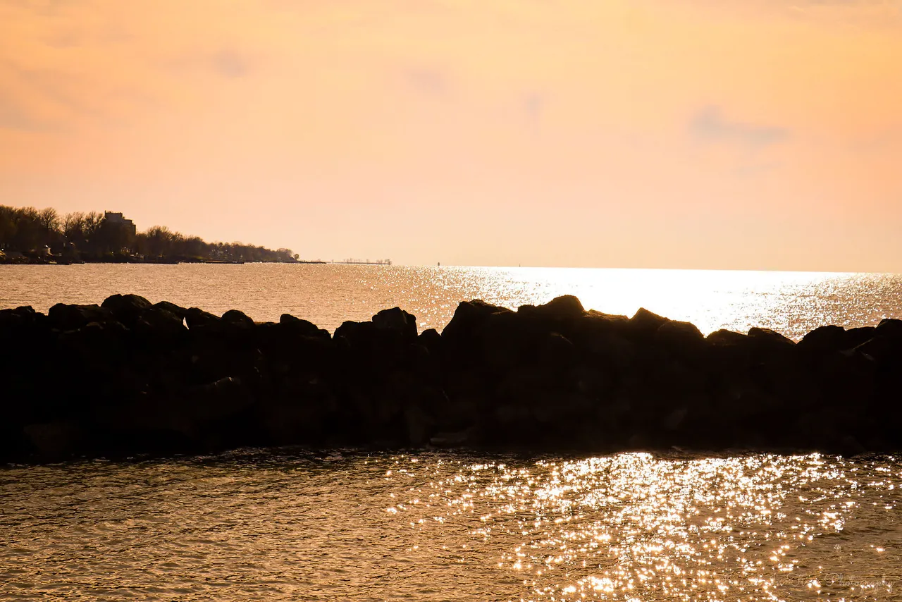 Dark jetty rocks against a golden evening sky, sunlight sparkling on the water.