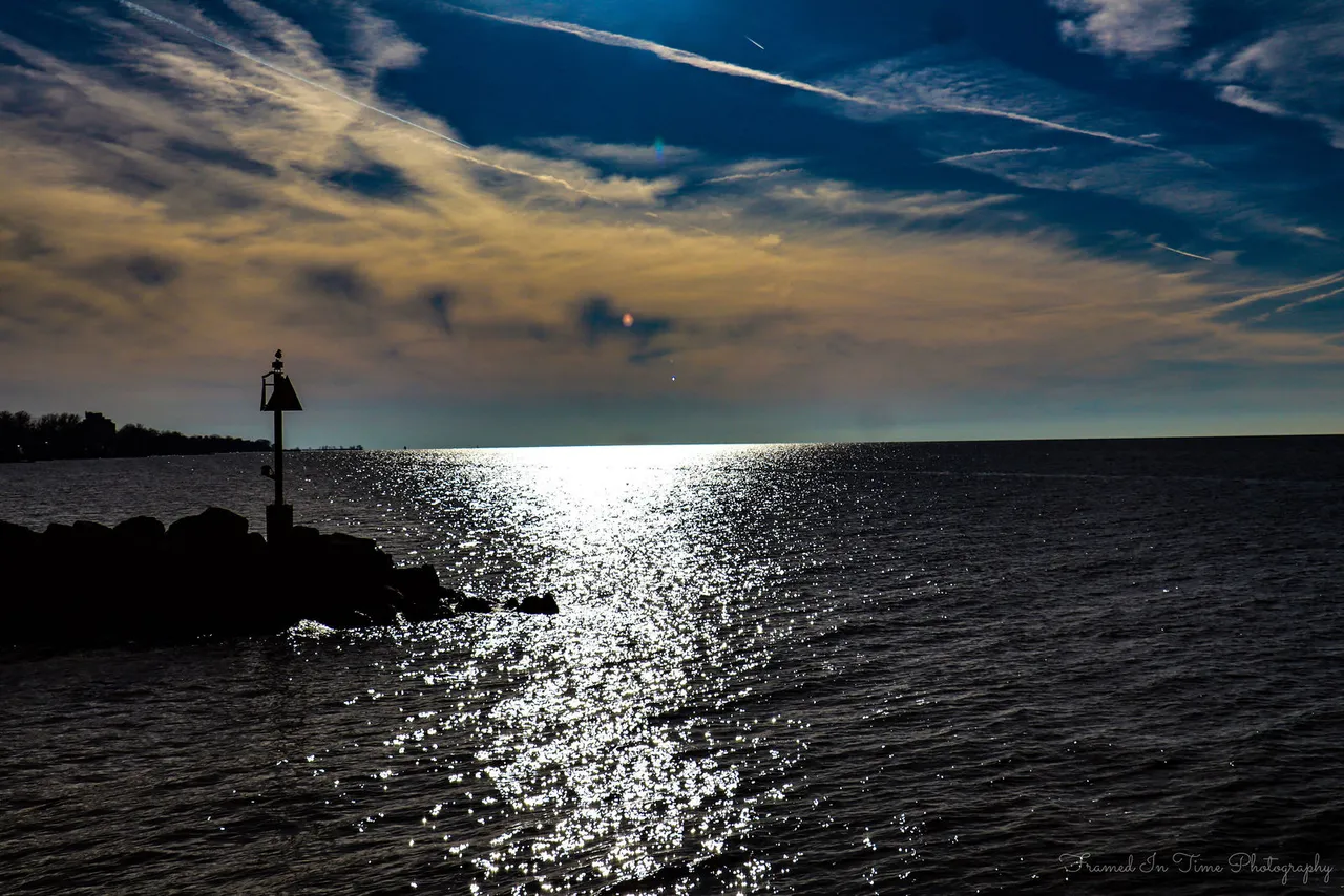 A jetty marker in silhouette on dark rocks, sunlight laying a glittering path across Lake Erie under streaked clouds.