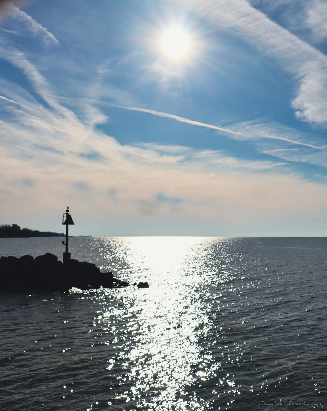 The sun high over Lake Erie, a path of light on the water running past a jetty marker in silhouette.