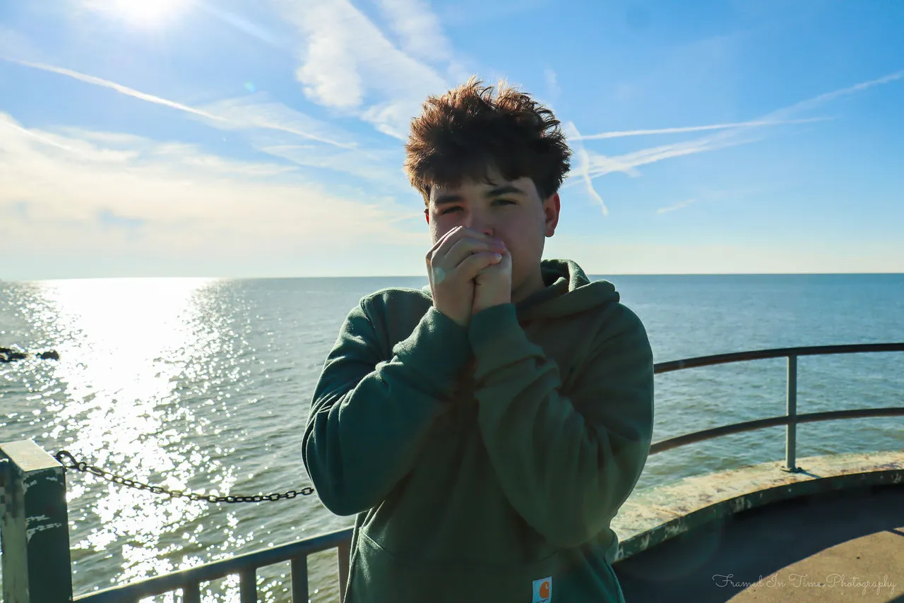 A boy in a green hoodie cupping his hands to his mouth against the wind on a sunny pier, open lake and streaked sky behind.