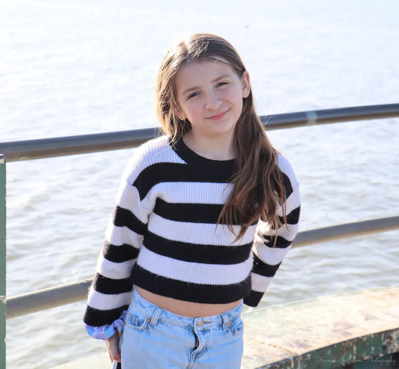 A girl with long brown hair in a black-and-white striped sweater, smiling with her head tilted, bright lake water behind her.