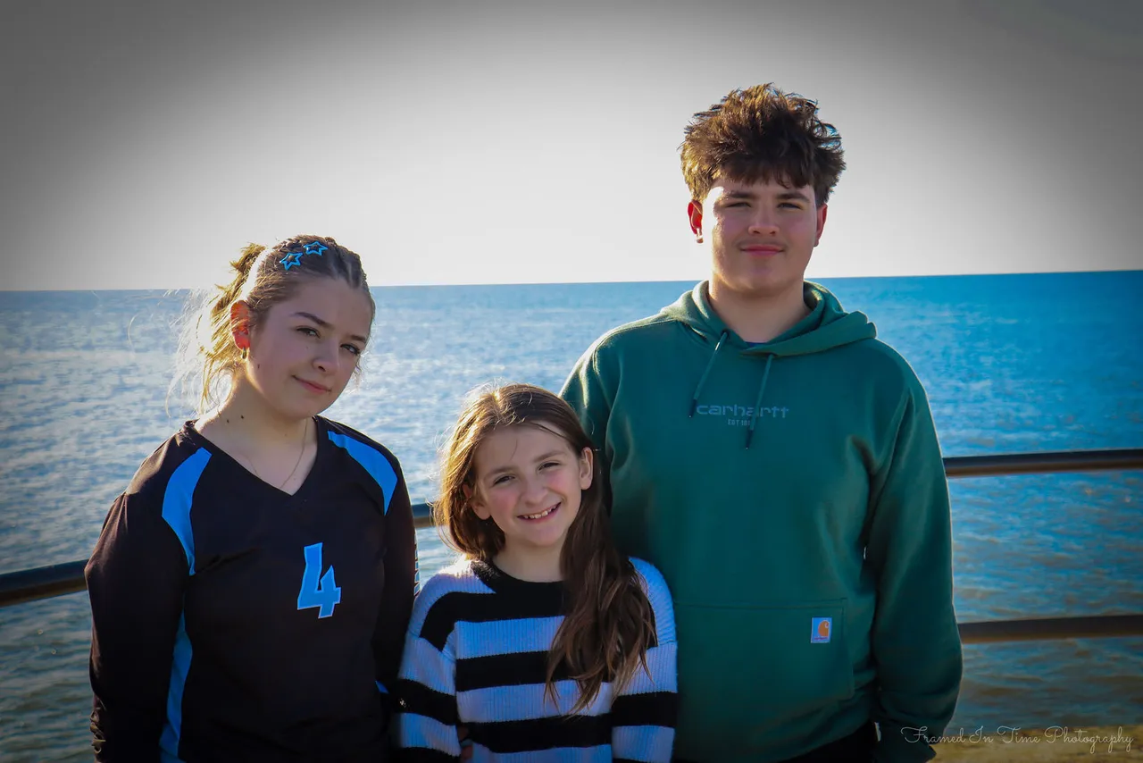 Three young people standing close together at a railing above the lake, the one in the middle grinning, blue water behind.